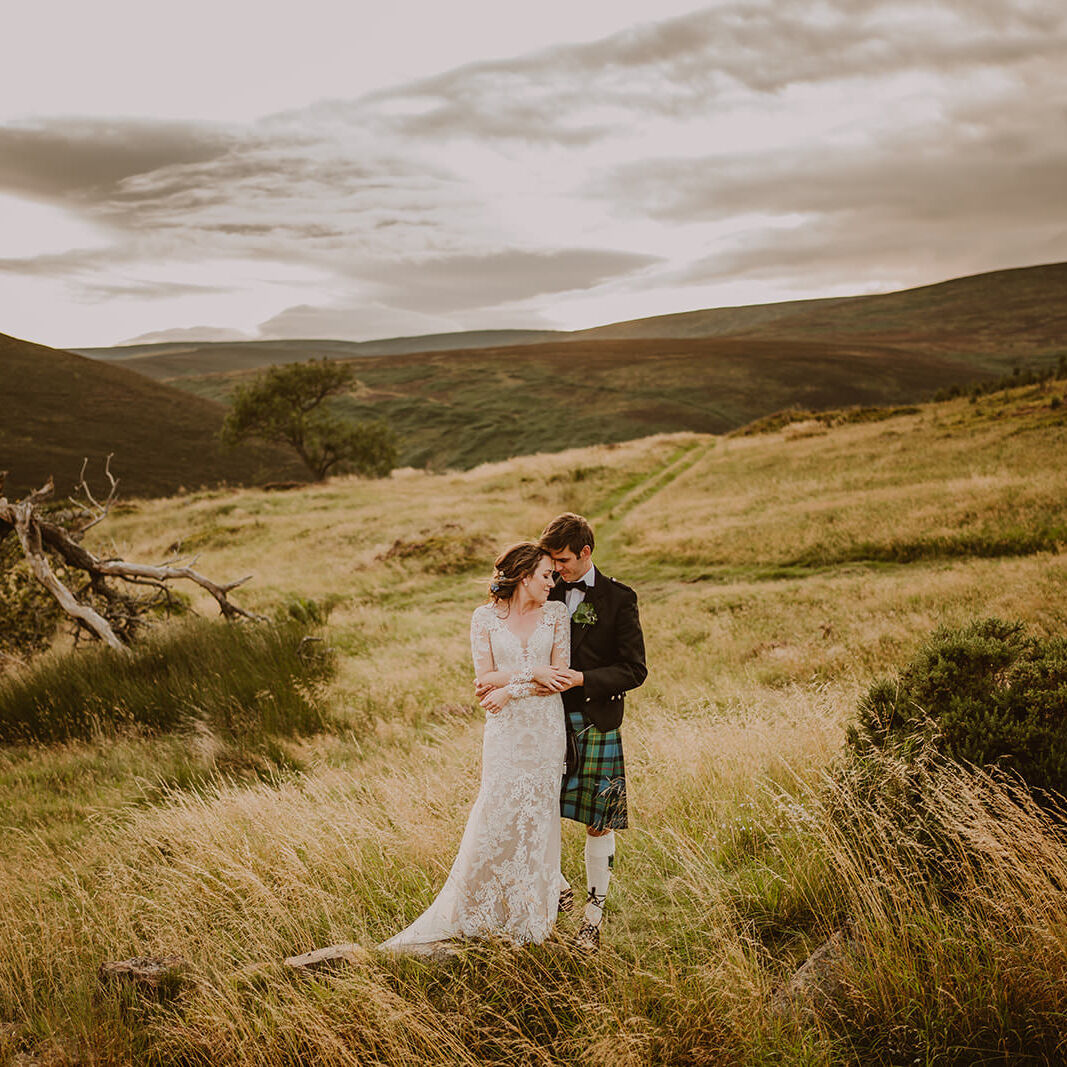 Couple standing together in long grass beneath the rolling hills of Scotland