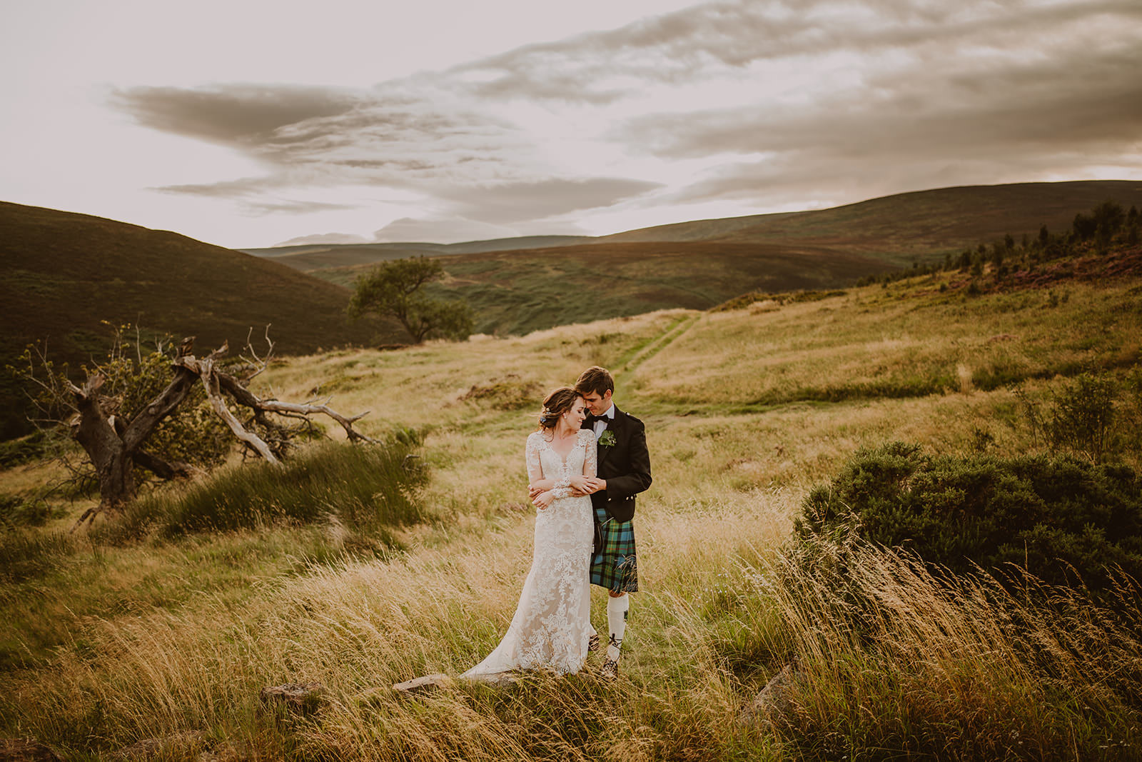 Couple standing together in long grass beneath the rolling hills of Scotland