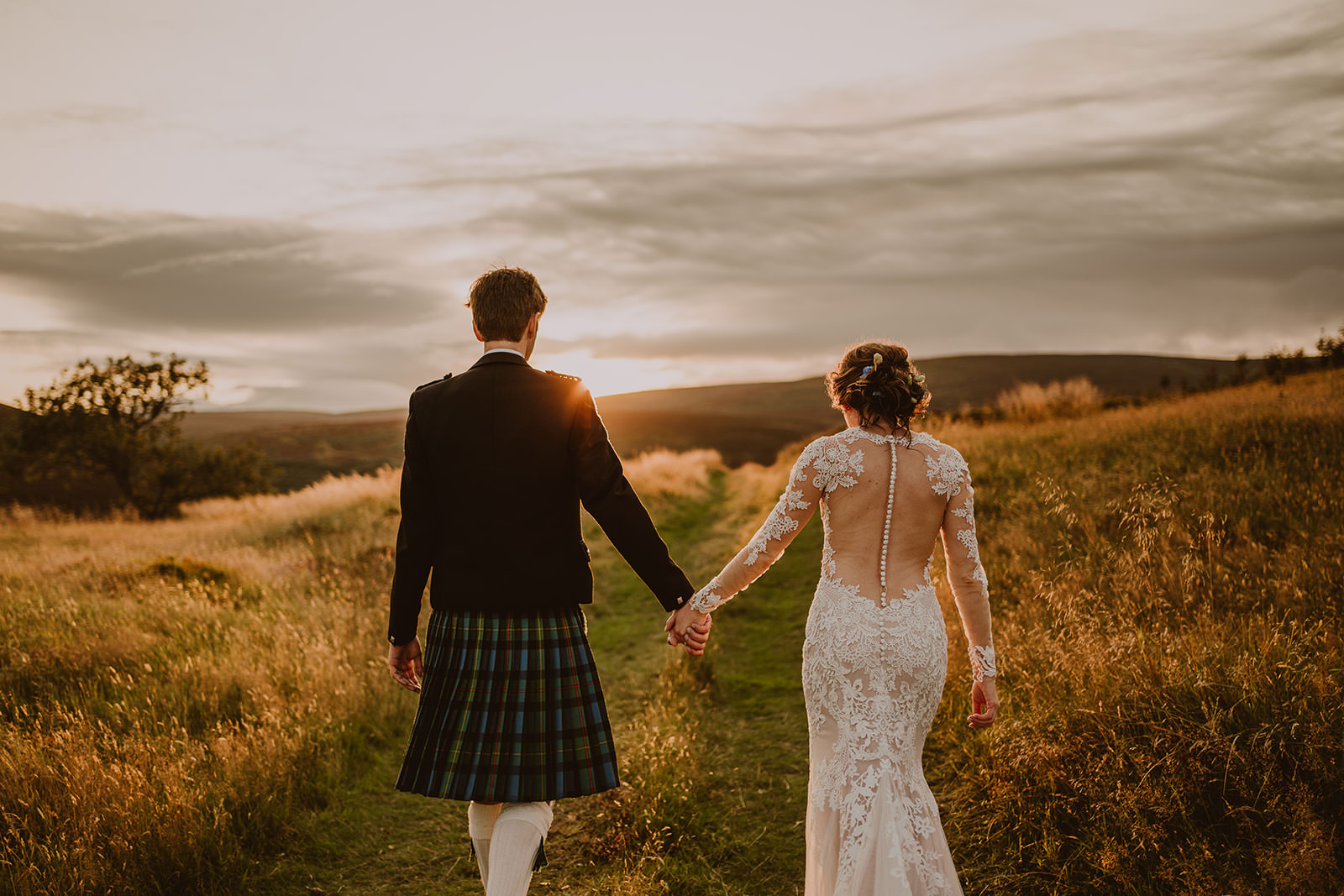 Newlyweds holding hands and walking through long grass at sunset